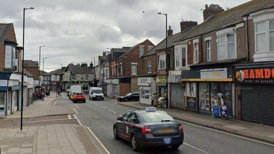 Streetview showing a grey taxi driving past a row of two-storey shops. One is a convenience store with the name "Local" written in large yellow letters on a yellow board above the door.