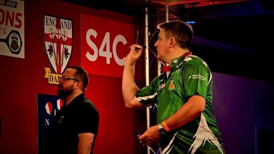 Llun o David Davies yng nghanol taflu dart. Mae'n gwisgo crys-t gwyrdd a gwyn. Mae ganddo wallt brown byr.