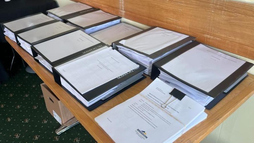 Nine folders of paperwork, plus a bundle of papers clipped together, on a wooden table. The green patterned carpet is visible on the floor, and a wood strip runs along the back wall of the room.