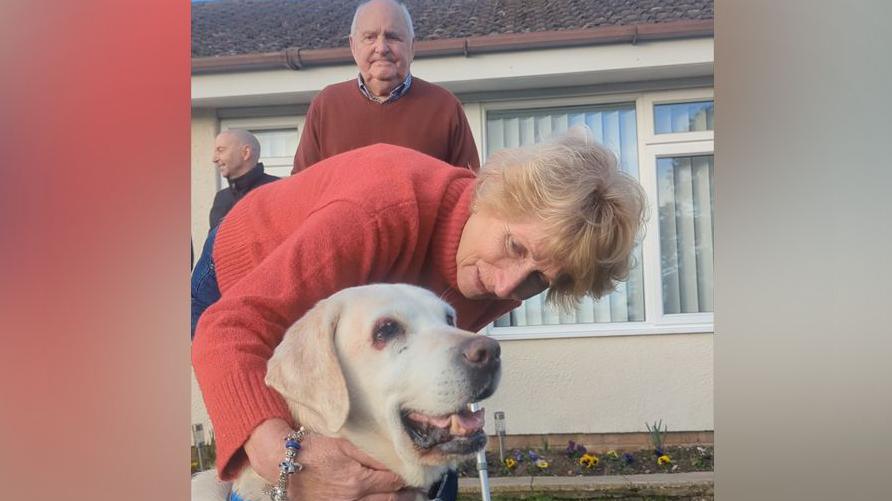 Rita hugging Daisy in her garden while Philip stands behind her, with a walking frame or stick. Rita wears a red/pink woollen roll neck jumper and blue jeans, and has a chunky bracelet on. Philip has a brown jumper and blue shirt on. Daisy's eyes look sore and her mouth is slightly open. She is a pale brown colour. 