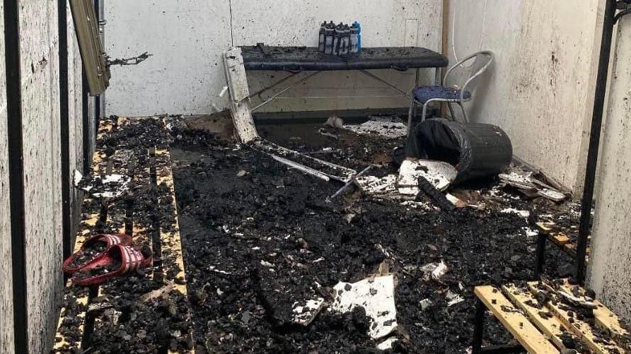 A picture showing the aftermath of the fire in the changing rooms. There is black burnt material covering the floor and benches. 