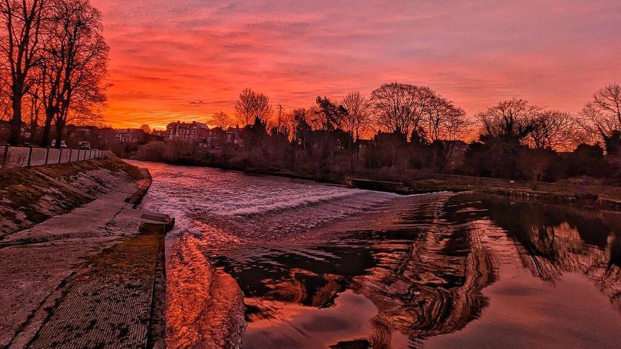 Red, yellow and purple colours can be seen in this view of the sky over a river weir. The water reflects the colours from the sunrise as it rolls over the weir. Trees can be seen outlined against the sky