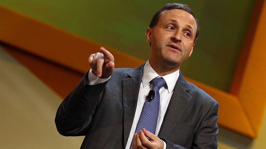 A man with short dark hair wearing a dark grey suit, purple tie and white shirt giving a presentation at a political conference. His right hand is raised with his first finger pointing at something off-camera.