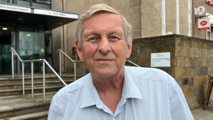 An man standing in front of some steps that lead to a council building. He has short light hair and is wearing a light blue and white check shirt, unbuttoned at the top.