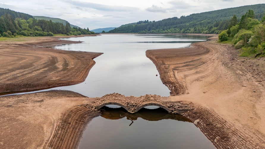 Stone bridge at the Llwyn-Onn Reservoir in Merthyr Tydfil in 2025. The bridge is visable above the water. A man is crossing the bridge with a fishing rod in his hand.