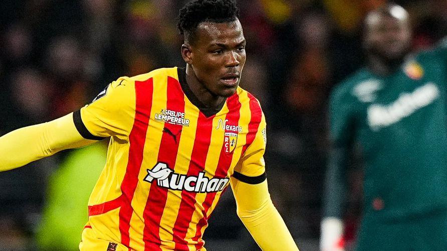 Juma Bah wears a red and yellow striped Lens shirt on top of a yellow undershirt which covers his arms during a football match as a goalkeeper is seen out of focus in the distance.