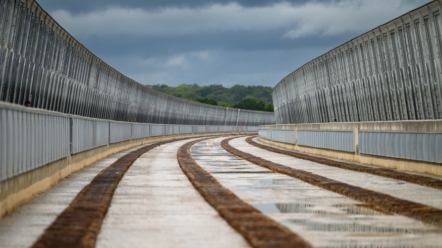 A bridge is curving to the right of the image and has silver barriers either side. There are brown raised pieces of material which runs across the bridge.