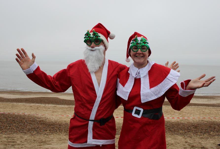 Two people dressed in Father Christmas outfits on Felixstowe beach. One is wearing a beard and the other is smiling at the camera