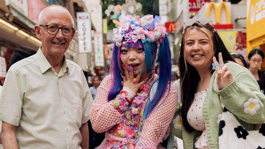 Colin and Charlotte in Japan with a Japanese woman standing between them dressed in pink net-sleeved top and purple hair in pig two pig tails with brightly coloured bows on top and purple eye shadow and nails.