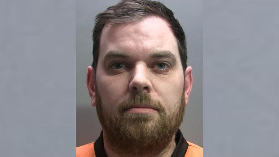 Mugshot of Andrew Bell who has a beard and short black hair. He is wearing an orange prison jumpsuit.