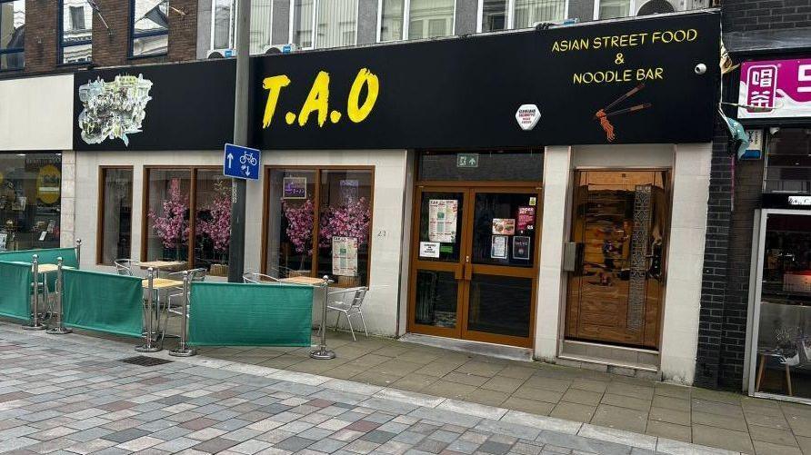 Front face of restaurant TAO. The signage is black with yellow print, and the window has pink flowers as decoration.  There are tables and chairs outside the site, which are empty.