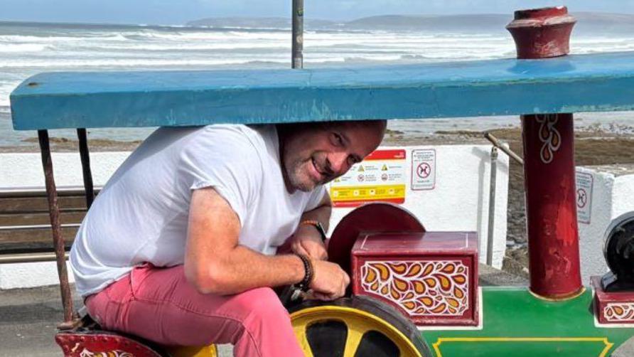 Adult Steve Dance is sitting on the seat of a toy steam engine. He has short brown hair and a beard and is wearing pink trousers and a white T-shirt with blue sandals. He is having to bend his head to fit under the canopy and looks too big for the ride. He is smiling and holding the steering wheel of the ride. It sits on the pavement and there is also an empty bench. The beach and sea are visible behind the pavement.