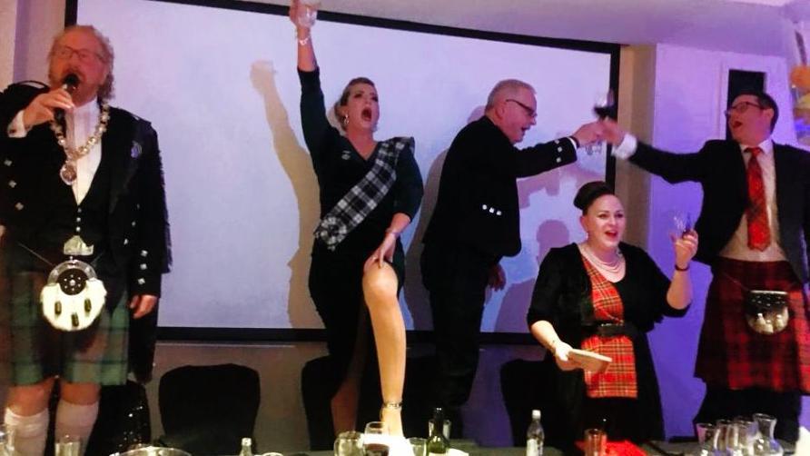 Five people stand at a table at a Burns supper, four with drinks in hand raised as if in toast. One woman, Kerry, has her leg on the table and has her glass held jovially in the air. 