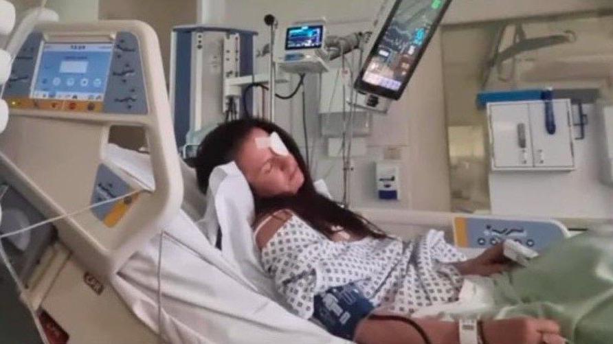 Kaylie lying on a hospital bed surrounded by machines. She has long brown hair and a white patch over her left eye. She is wearing a white gown and her head is at an angle suggesting she is asleep.