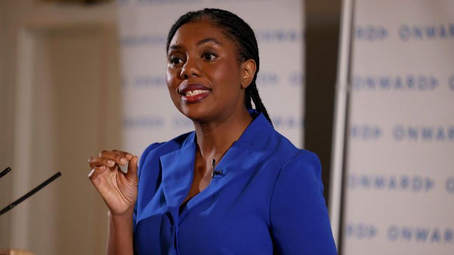 Conservative Party Leader Kemi Badenoch, wearing a blue dress, gesticulates to punctuate her speech from behind a lectern on how the Conservative party can rebuild voter