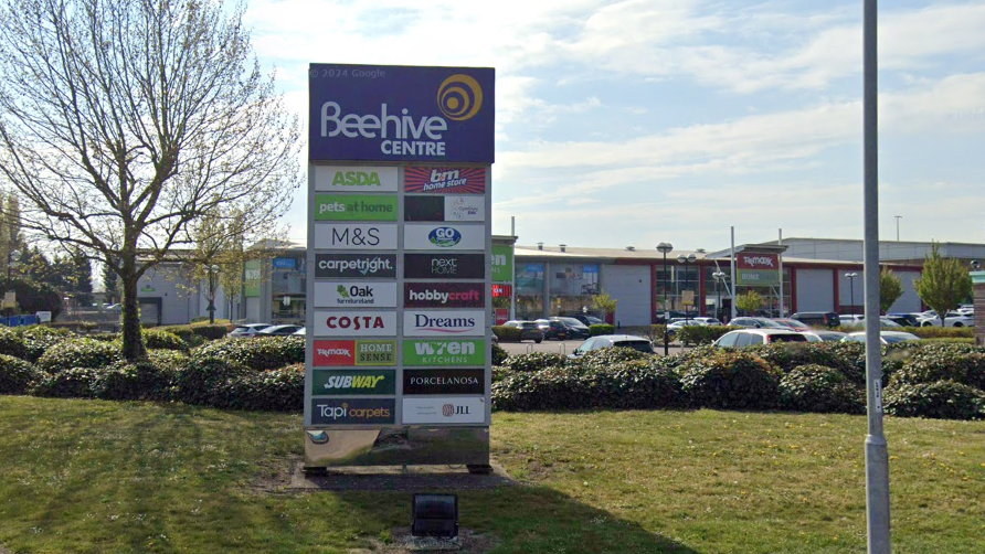 A sign saying 'Beehive Centre' with various shop names underneath.
It is erected on green grass and a tree on the left hand side.