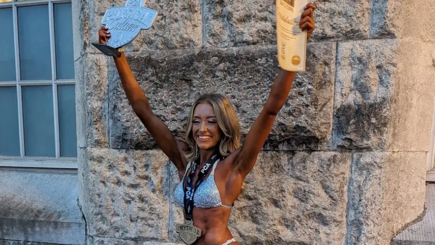 Mia Collins wearing a silver bikini and with a dark spray tan. She is standing with her arms in the air holding awards and wearing a medal round her neck.