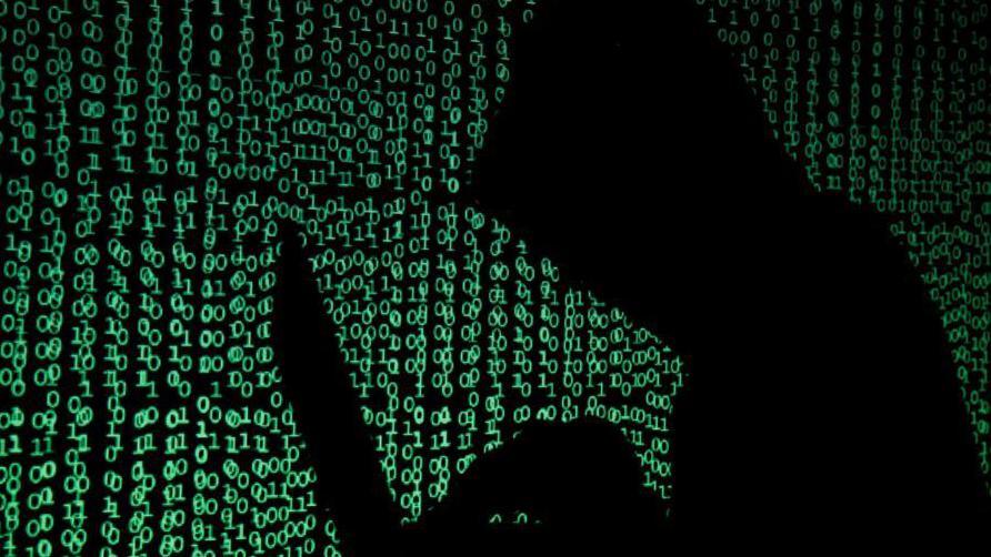 A hooded figure in shadow is working at a laptop in front of a background with green letters on it.