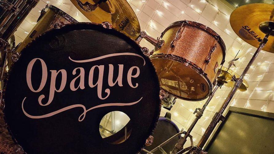 A drum kit in front of a white tiled wall. One of the drums is black with Opaque written in pink letters on the front. 