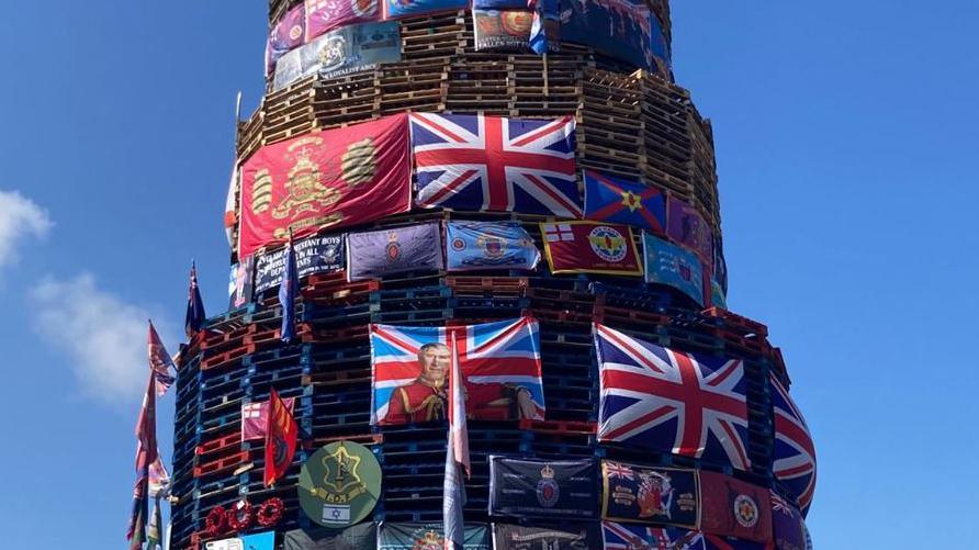 A large number of flags including union flags and a flag depicting an image of King Charles placed on a bonfire in Derry's Bogside.