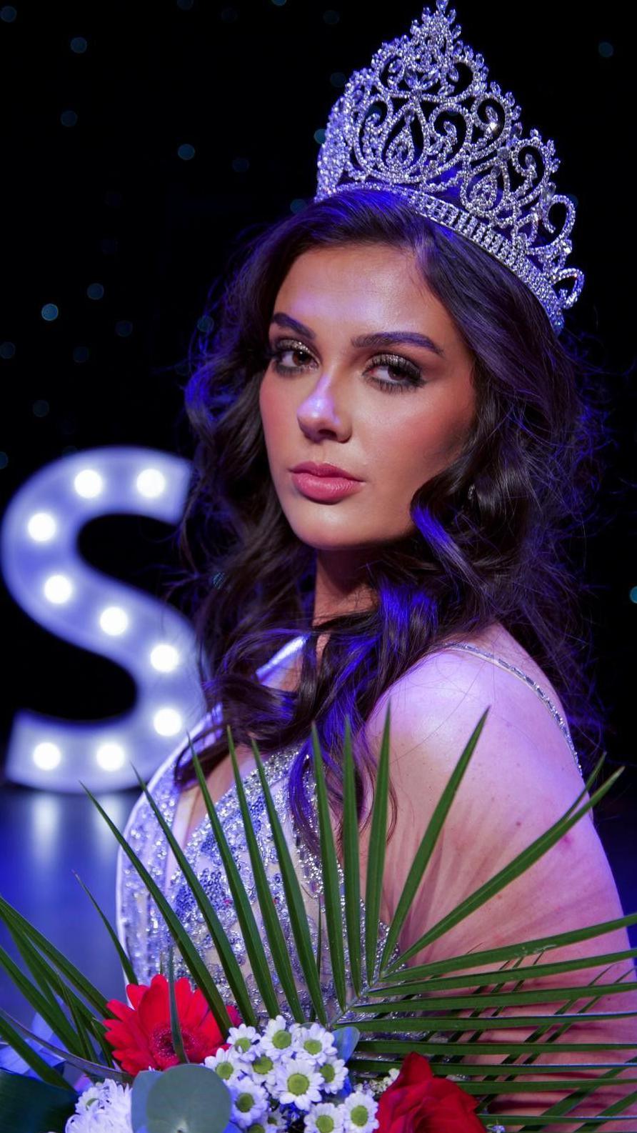 A woman with long dark wavy hair, wearing a crown and a sparkly gown, holding a bouquet of flowers