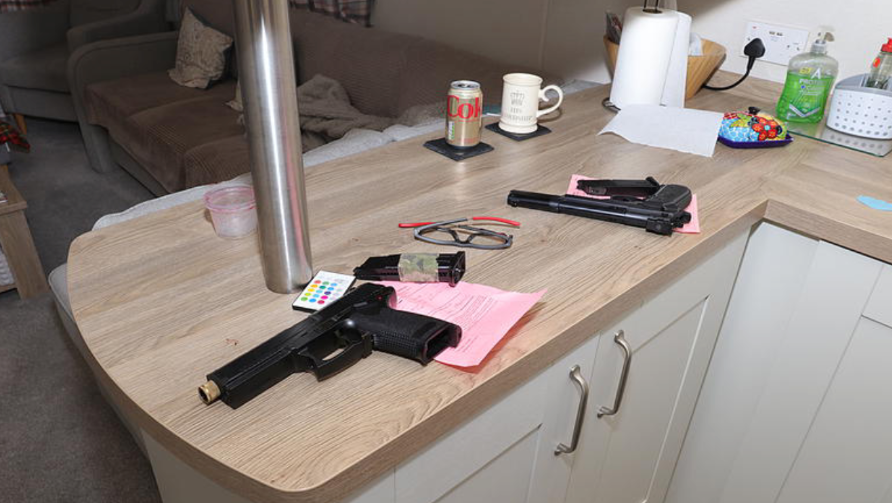 Two black guns lie on a kitchen counter-top. The magazine has been taken out and sits beside each gun. Behind the guns on the counter, a Coke can, a coffee mug and a roll of kitchen can be seen; a settee is visible in the background.