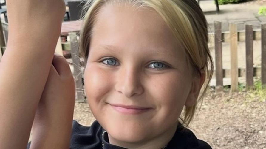 A smiling girl with blue eyes and fair hair pulled back into a ponytail. She is wearing a black top. She is outdoors and there is a wooden fence behind her.