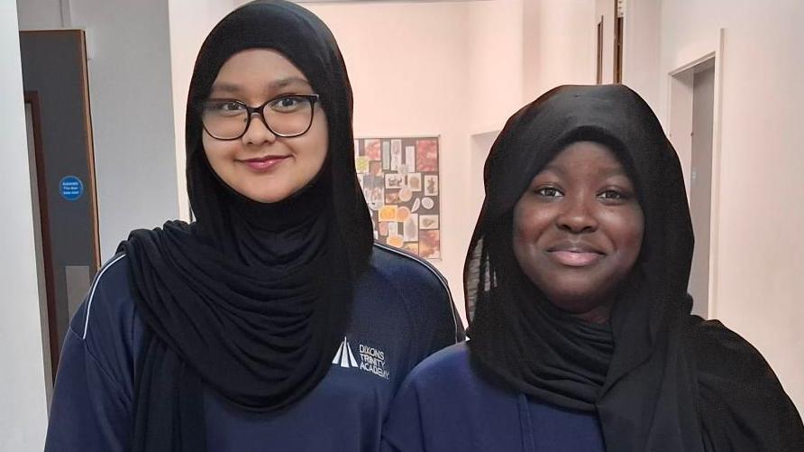 Two girls stand alongside each other in a school corridor. They are wearing blue sweatshirts that have the Dixons Trinity Academy logo on them. The girl on the left is wearing glasses and a black headscarf. The girl on the right is also wearing a black headscarf.