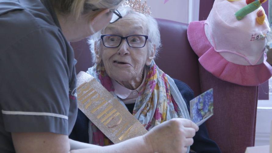 An elderly man in a birthday sash is shown a card by a nurse.