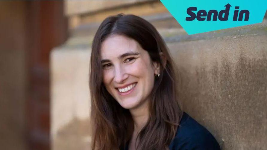 Katherine Rundell smiling leaning against a building. She has long dark hair and is wearing a navy coloured top.