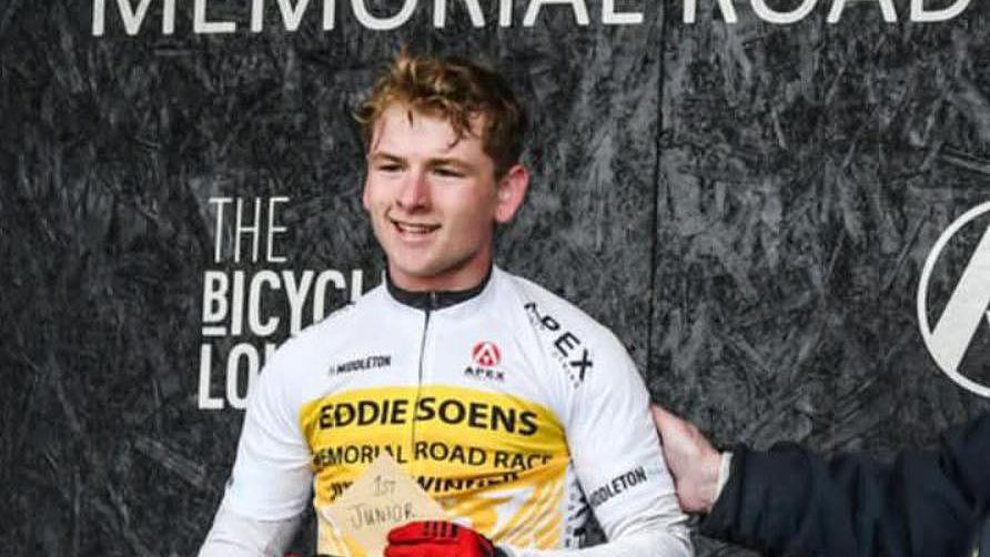A young man - Aidan Worden - smiles, holding a piece of paper that reads '1st Junior'. He wears a white cycling jersey and red gloves.