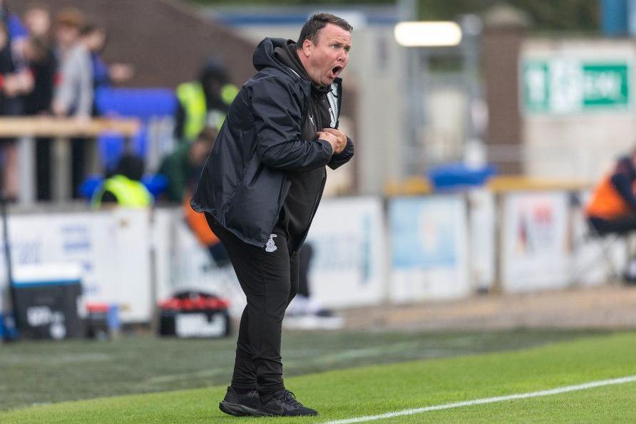 Scott Kellacher is dressed in black football training kit and a black waterproof jacket. He is standing at the byline of a football pitch, shouting instructions to football players, who are out of shot. Out of focus behind Kellacher is a stand of the football ground with some spectators.