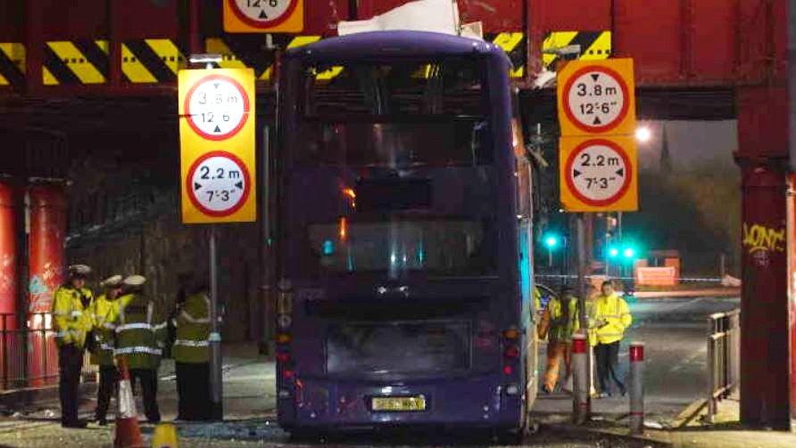 The rear view of a bus wedged under a railway bridge surrounded by police officers. The area is taped off