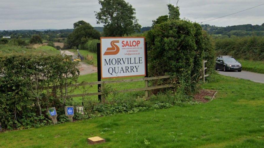 A sign reading "Morville Quarry" under a brown logo for the site owners Salop Sand and Gravel. The sign sits atop a wooden fence on a grass verge with hedges either side and an access road visible behind.