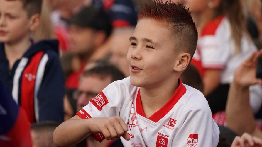 A young fan with red spikey hair and wearing a red and white shirt is smiling in a crowd of people.