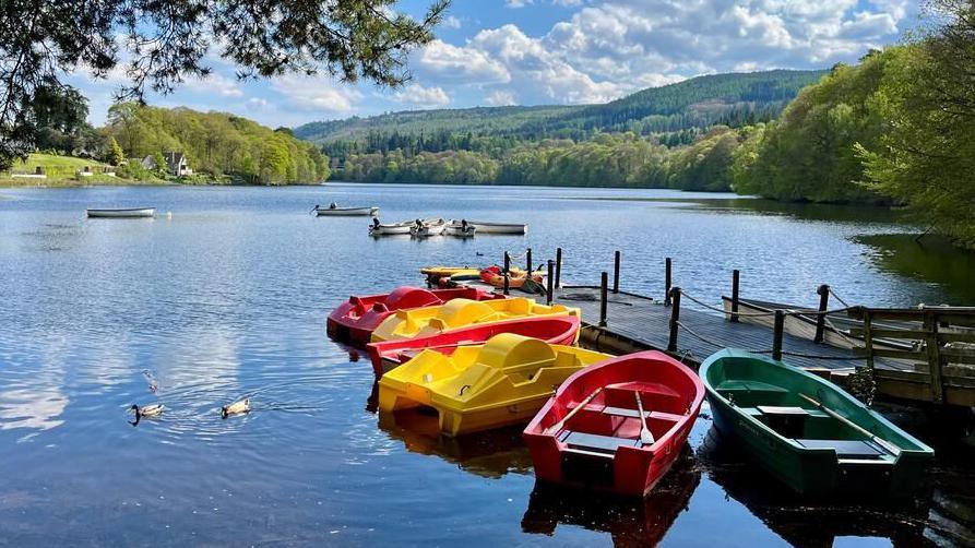 A loch with a pier and colourful plastic pedal boats floating in the water. Green hills and trees can be seen in the distance and the sky is blue with some clouds