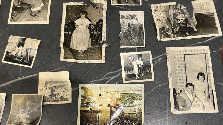 Family photos are spread out across a kitchen table, showing black and white images of children and families as well as a few colour images.