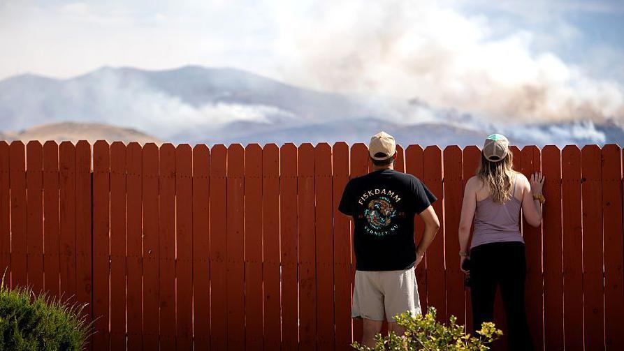 Two people look over what appears to be a domestic fence. They are looking at where smoke is rising from hills in the background. 
