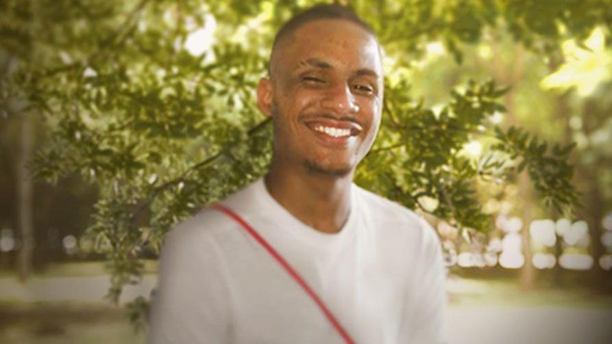 A smiling 20-year-old man in a white t-shirt standing in a park