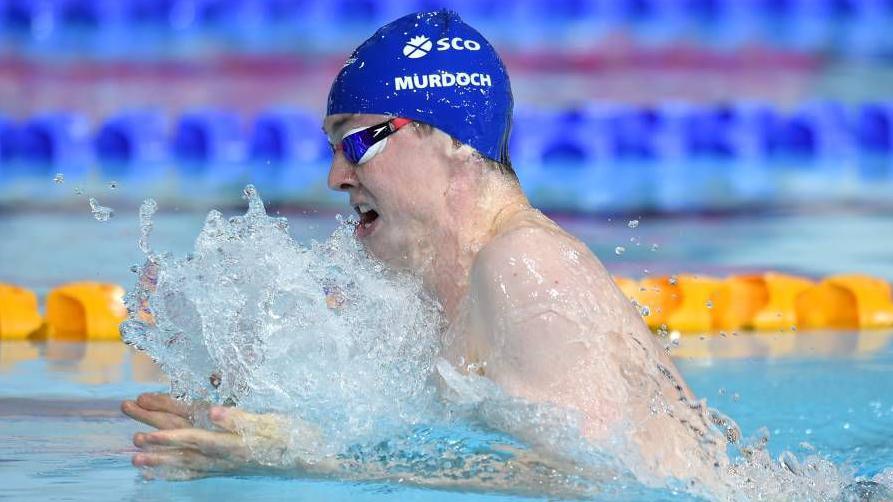 Team Scotland athlete Ross Murdoch mid breast stroke during Glasgow 2014. He is wearing a dark blue swimming cap and goggles.