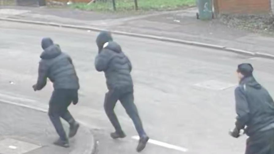 Three men, all wearing black clothes, run along a residential street. They all have their backs to the camera.