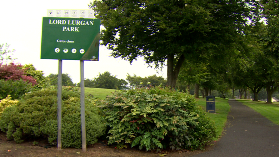 A sign saying Lord Lurgan Park. There are bushes, and trees in view and a pathway.