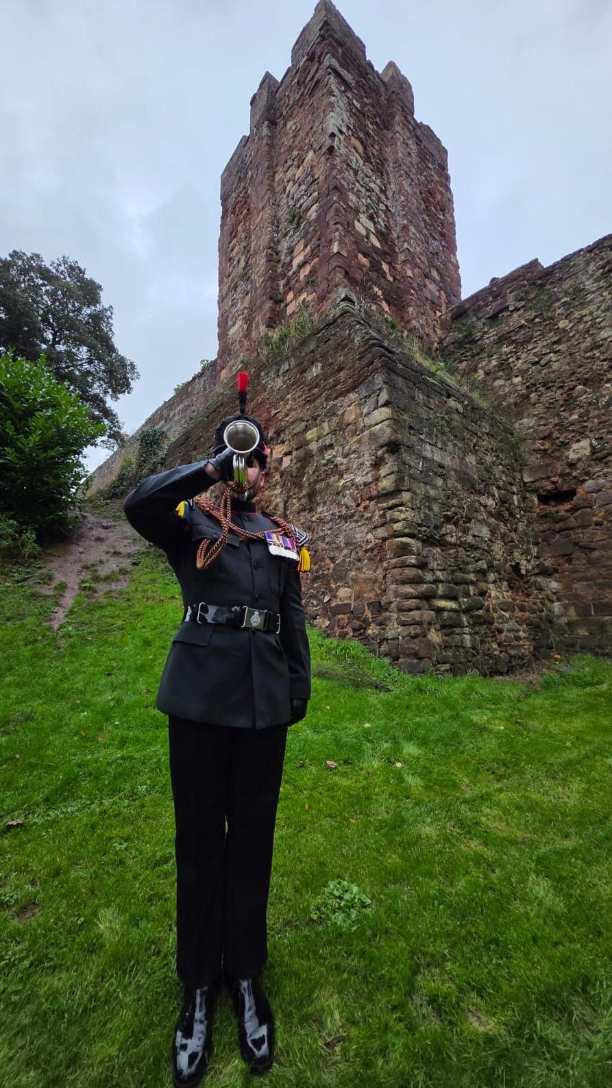 A man in a military uniform plays the bugle. He is standing up straight. Behind him is a stone structure.