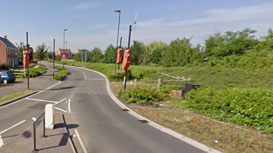 A suburban road on a sunny day. New build red brick houses are visible on the left, while green scrubland sits to the right of the road. A number of traffic lights in red covers are also visible. 