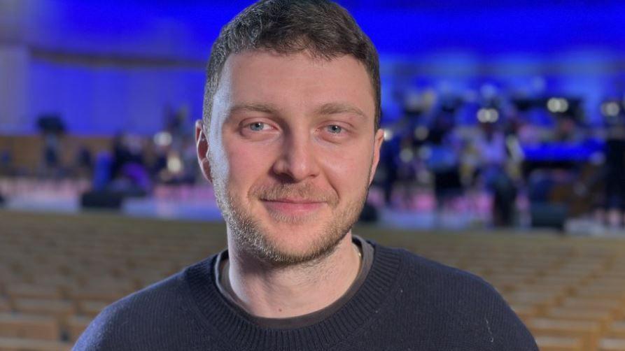 Head and shoulders shot of Adam Wood smiling at the camera. The blurred background appears to be an orchestra.