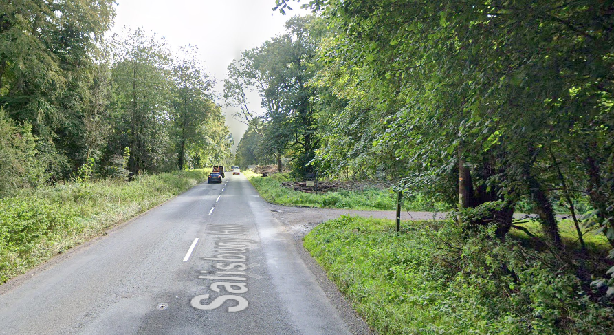 A main road through a forested area with a turning on the right and cars both approaching and driving away from the camera