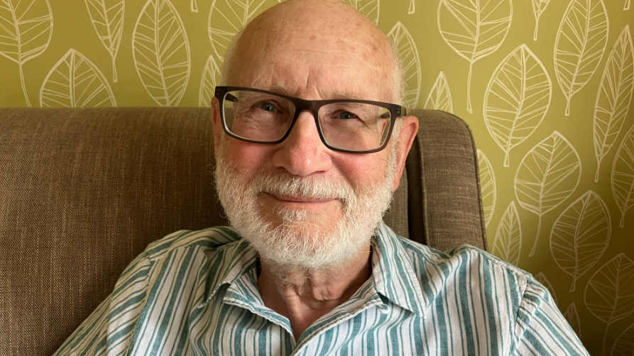 Malcolm Pemble sitting in an armchair is wearing a striped shirt, and dark framed glasses. He has blue eyes and a white beard.
