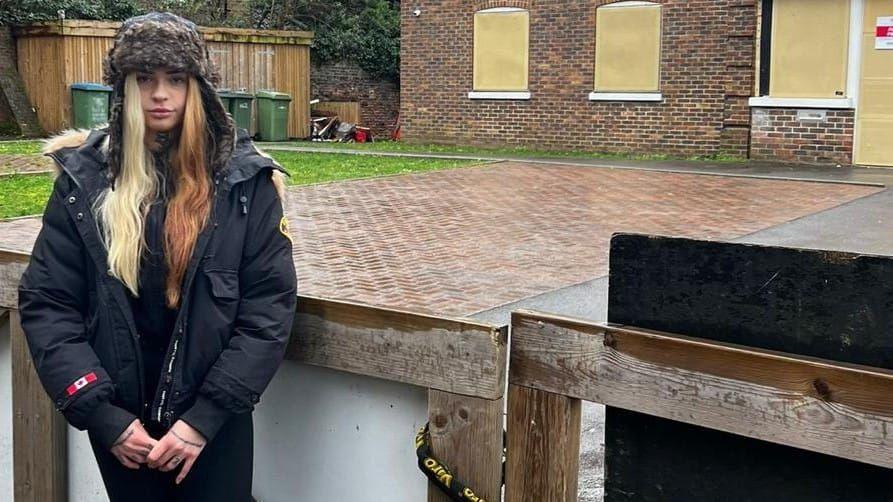 A young woman stands in front of a padlocked gate and boarded up building