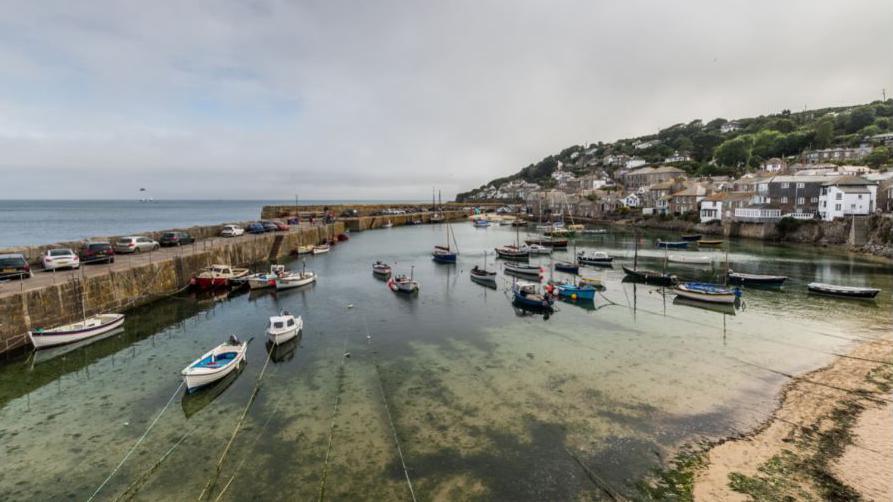 Small boats are moored in a harbour at low tide. One one side of the harbour there is a sea wall, with some cars parked on it. An opening is just visible towards the far right. There are houses and other low level buildings in  the top right hand side of the image and they are nestled besides the sea. There is a slight hill above them. Seaweed can be seen through the clear water in the harbour.