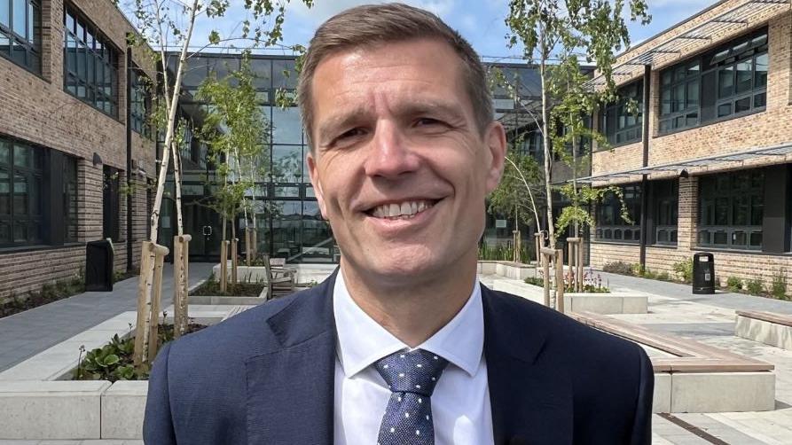 The head teacher of Bishop's Stortford High School, Dale Reeve. He has short, brown hair and is wearing a blue jacket, shirt and tie. He is smiling at the camera. Part of the school, a two-storey modern building, can be seen in the background.